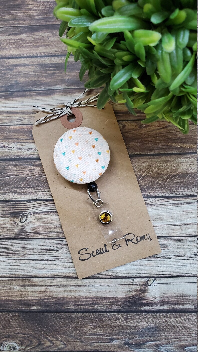 May include: A white fabric badge reel with a colorful heart pattern. The reel has a black retractable cord and a clear plastic badge holder with a gold-colored button. The reel is attached to a brown card with the text "Scout & Remy" written in black.