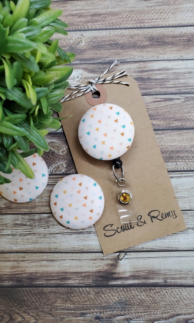 May include: A white fabric button with a pattern of small orange, teal, and white hearts. The button is attached to a retractable badge holder with a clear plastic case and a gold-colored clasp. The badge holder is attached to a brown card with the text "Scotti & Remy".