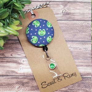 Puede incluir: Un portagafete retr&aacute;ctil de tela azul y verde con un patr&oacute;n de rana y corazones rosas. El portagafete est&aacute; unido a una tarjeta marr&oacute;n con el texto "Scout & Remy".