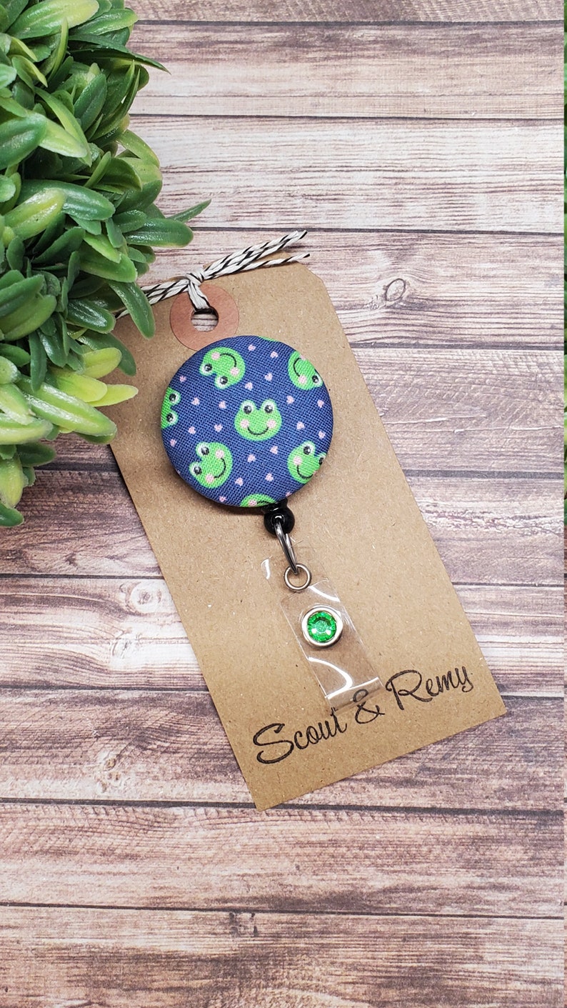 Puede incluir: Un portagafete retr&aacute;ctil de tela azul y verde con un patr&oacute;n de rana y corazones rosas. El portagafete est&aacute; unido a una tarjeta marr&oacute;n con el texto "Scout & Remy".