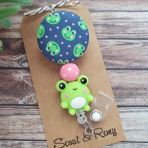 May include: A green frog-shaped badge reel with a white belly and black eyes, attached to a clear plastic holder. The reel features a navy blue fabric button with green frog faces and pink hearts. The tag reads "Scout & Remy".
