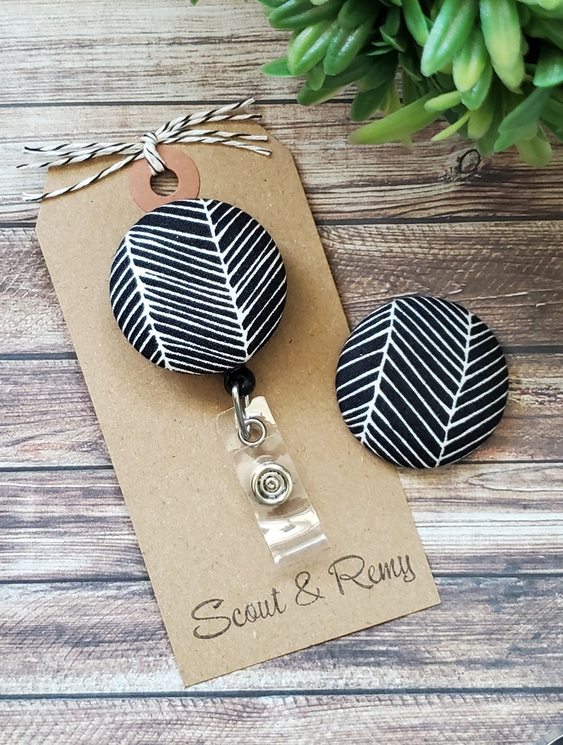 May include: Black and white patterned fabric badge reel with a clear plastic retractable cord and a silver clip. The badge reel is attached to a brown card with the text "Scout & Remy" printed on it.