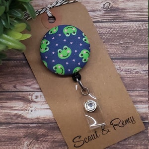 Puede incluir: Un portagafete retr&aacute;ctil de tem&aacute;tica de rana azul y verde con un portagafete de pl&aacute;stico transparente y un cierre plateado. El portagafete est&aacute; unido a una tarjeta marr&oacute;n con el texto "Scout & Remy".