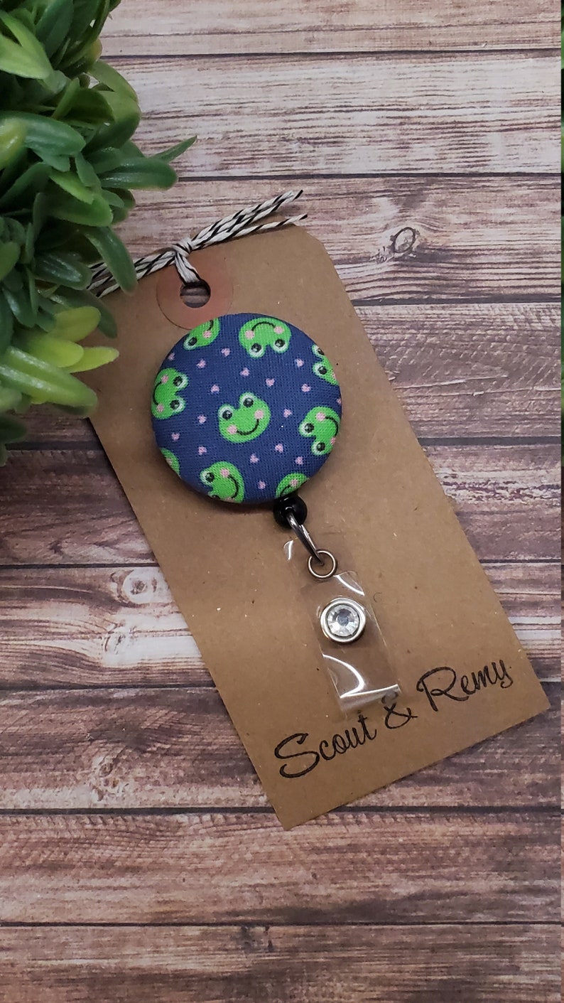 Puede incluir: Un portagafete retr&aacute;ctil de tem&aacute;tica de rana azul y verde con un portagafete de pl&aacute;stico transparente y un cierre plateado. El portagafete est&aacute; unido a una tarjeta marr&oacute;n con el texto "Scout & Remy".