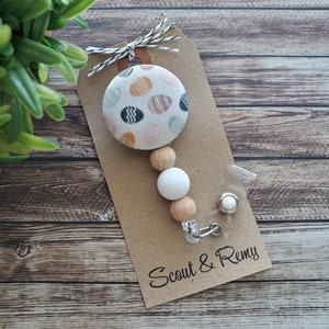 May include: A brown paper tag with the text "Scout & Remy" printed on it. A white and brown beaded lanyard with a white button featuring a colorful polka dot pattern is attached to the tag.
