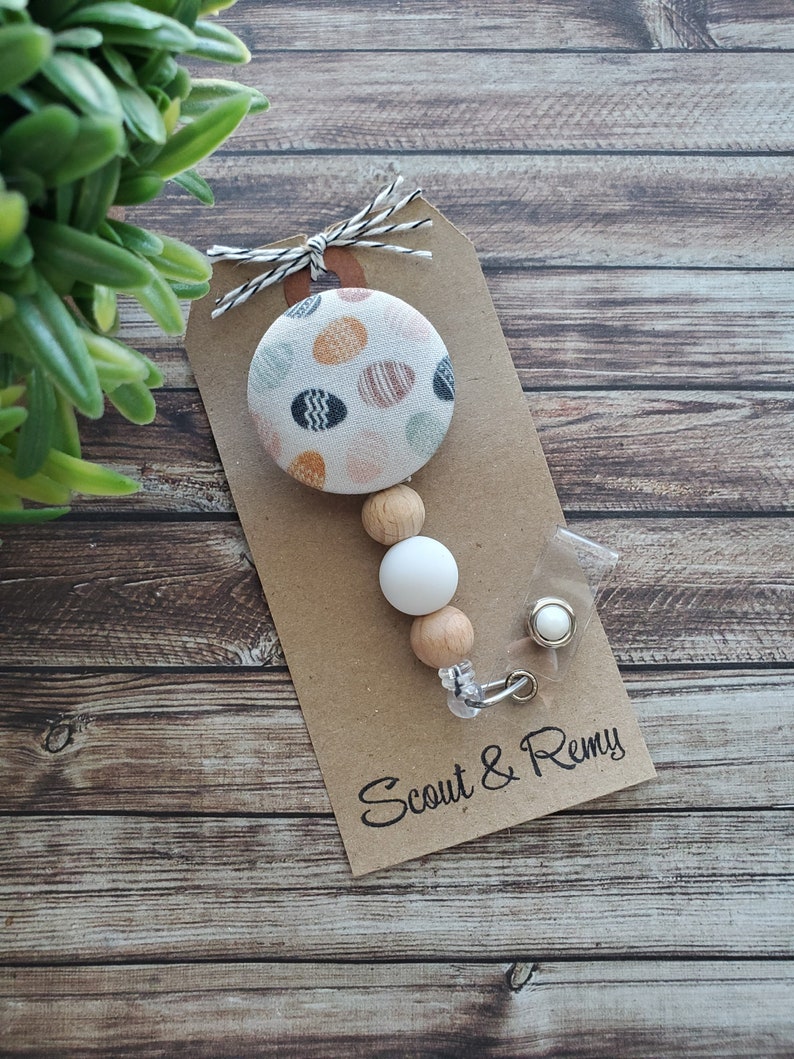 May include: A brown paper tag with the text "Scout & Remy" printed on it. A white and brown beaded lanyard with a white button featuring a colorful polka dot pattern is attached to the tag.