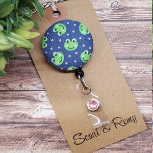 Puede incluir: Un carrete de insignia de tela azul con un patr&oacute;n de rana verde y corazones rosas. El carrete tiene un cord&oacute;n retr&aacute;ctil negro y un porta insignias de pl&aacute;stico transparente con un strass rosa. La tarjeta adjunta al carrete dice "Scout & Remy".