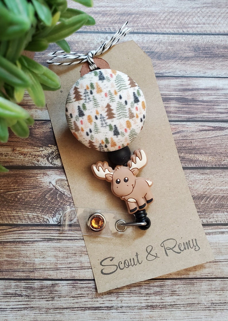 Pu&ograve; includere: Un portabadge con un bottone circolare in tessuto con motivo a alberi di bosco multicolore, fissato a un ciondolo in silicone a forma di alce marrone. Il portabadge include un cinturino in plastica trasparente con bottone a pressione e strass.