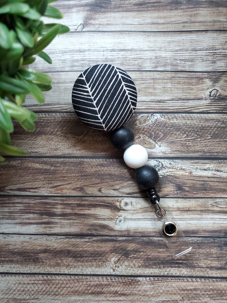 May include: Black and white patterned fabric badge reel with black and white beads. The badge reel has a clear plastic strap and a metal clip.