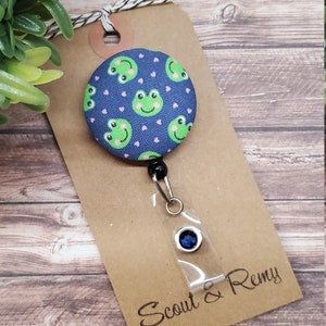 Puede incluir: Un portagafete retr&aacute;ctil de tem&aacute;tica de rana azul y verde con un strass azul. El portagafete est&aacute; unido a una etiqueta marr&oacute;n con el texto "Scout & Remy".