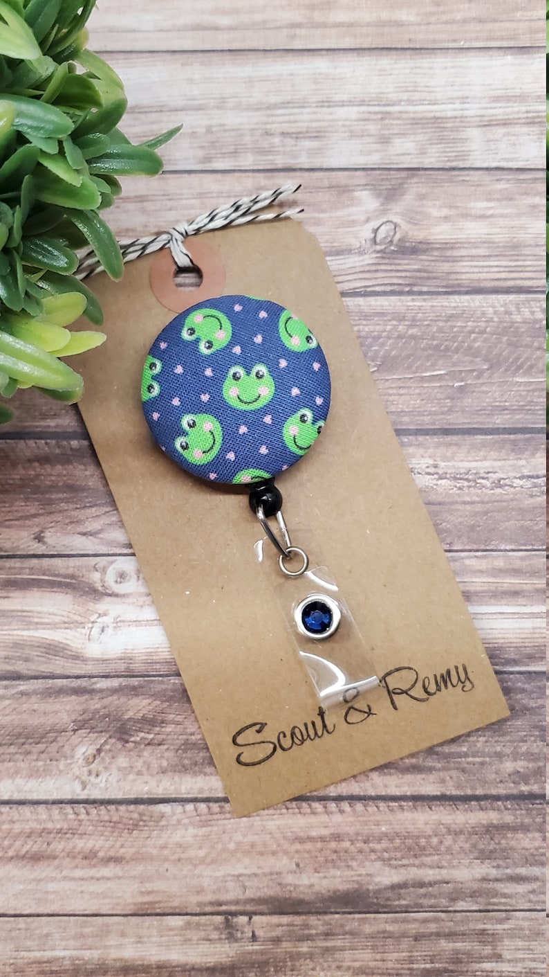 Puede incluir: Un portagafete retr&aacute;ctil de tem&aacute;tica de rana azul y verde con un strass azul. El portagafete est&aacute; unido a una etiqueta marr&oacute;n con el texto "Scout & Remy".