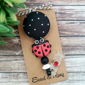 May include: A ladybug-themed badge reel with a black polka-dot top and a red and black ladybug design. The reel includes a clear plastic clip and a red gem accent. The card reads "Scout & Remy".