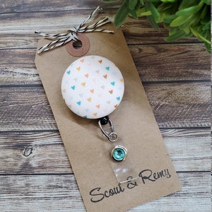 May include: A white fabric badge reel with a button featuring a pattern of small orange, pink, and blue hearts. The reel has a clear plastic badge holder with a turquoise rhinestone. The tag is brown with the text "Scout & Remy" in black.