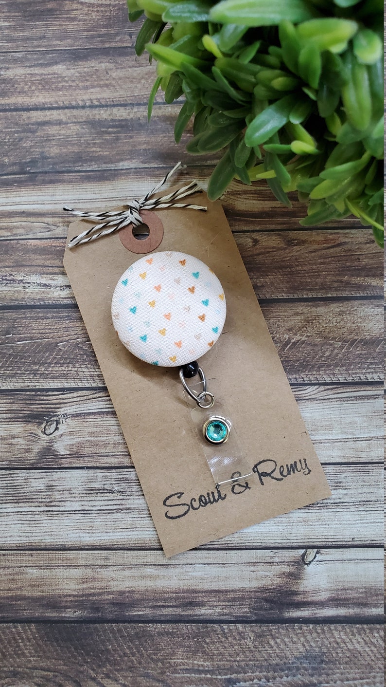 May include: A white fabric badge reel with a button featuring a pattern of small orange, pink, and blue hearts. The reel has a clear plastic badge holder with a turquoise rhinestone. The tag is brown with the text "Scout & Remy" in black.