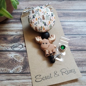 Pu&ograve; includere: Un portabadge con un bottone circolare in tessuto decorato con un motivo boschivo di alberi verdi, arancioni e marroni. Sotto il bottone si trova un ciondolo in silicone a forma di alce marrone, una perlina nera e una clip in plastica trasparente.