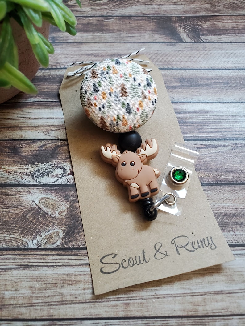 Pu&ograve; includere: Un portabadge con un bottone circolare in tessuto decorato con un motivo boschivo di alberi verdi, arancioni e marroni. Sotto il bottone si trova un ciondolo in silicone a forma di alce marrone, una perlina nera e una clip in plastica trasparente.