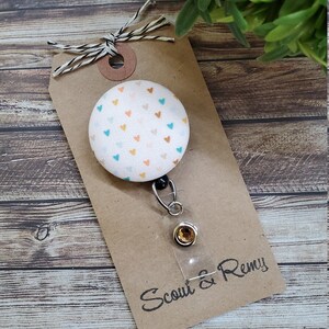 May include: A white fabric badge reel with a colorful heart pattern. The reel has a clear plastic badge holder and a silver retractable cord. The reel is attached to a brown card with the text "Scout & Remy" in black.