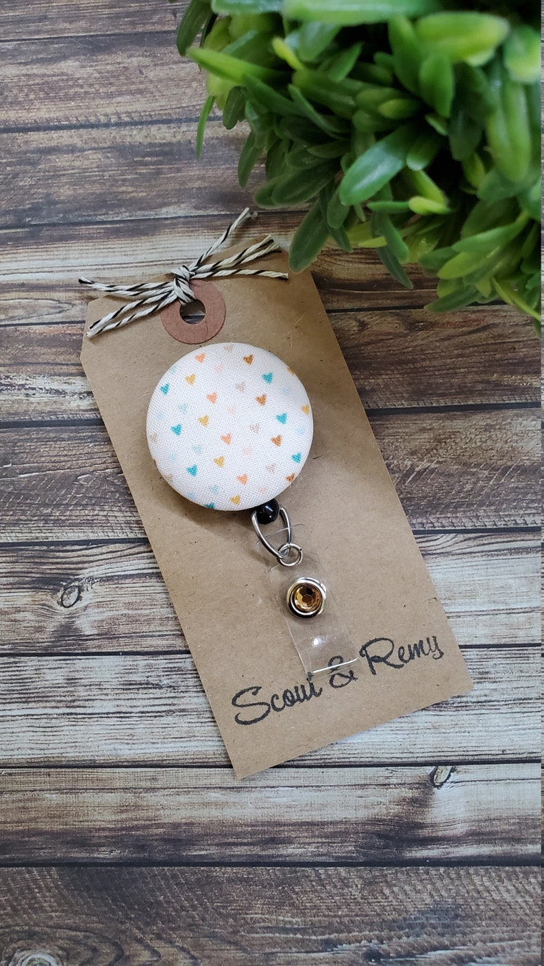 May include: A white fabric badge reel with a colorful heart pattern. The reel has a clear plastic badge holder and a silver retractable cord. The reel is attached to a brown card with the text "Scout & Remy" in black.