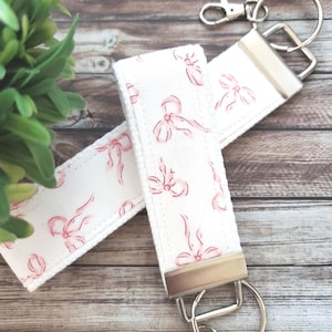 May include: Two white key fobs with a pink bow pattern. Each fob has a silver-colored metal clasp and a split ring. The key fobs are lying on a wooden surface, with green foliage in the background.