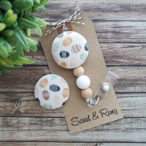 May include: A white fabric button with a colorful Easter egg pattern, a wooden bead necklace with a white bead, and a clear plastic badge holder with a silver clip. The tag reads "Scout & Remy".