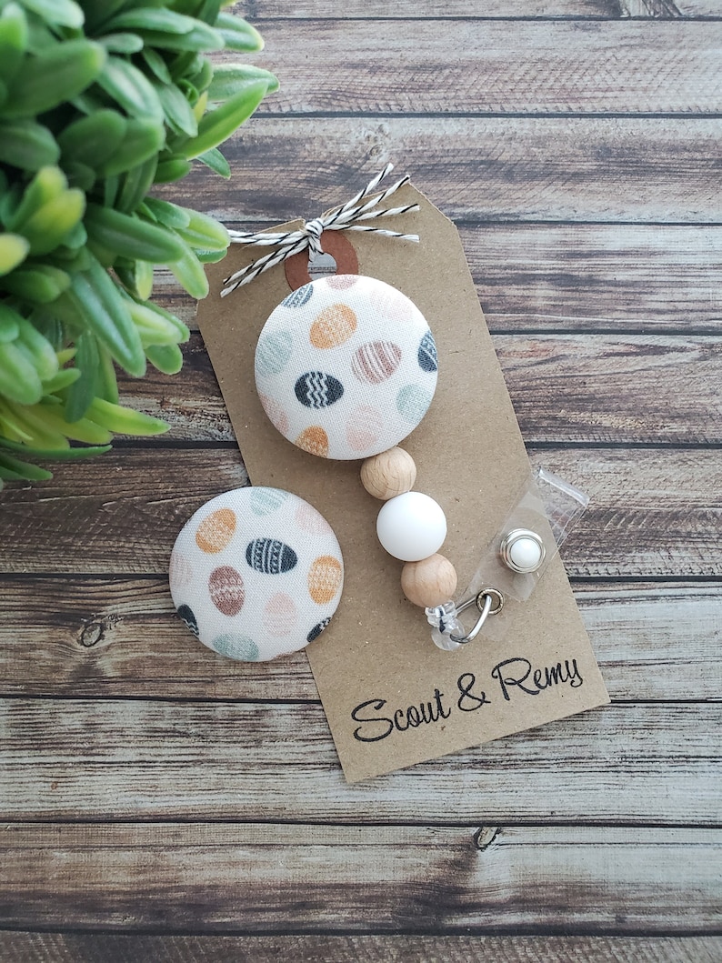 May include: A white fabric button with a colorful Easter egg pattern, a wooden bead necklace with a white bead, and a clear plastic badge holder with a silver clip. The tag reads "Scout & Remy".