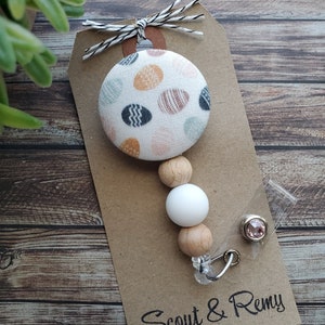 May include: A white fabric button with a colorful geometric pattern, attached to a wooden bead and white silicone bead, with a clear badge holder and a pink rhinestone. The tag reads "Scout & Remy".