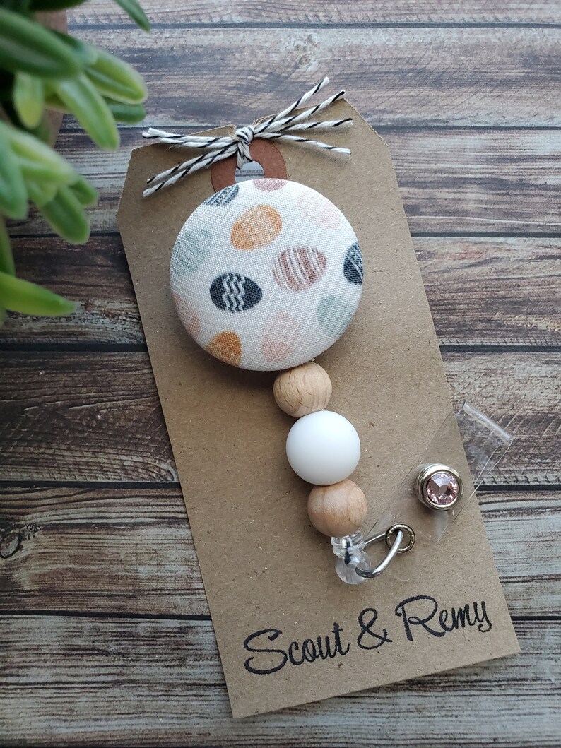 May include: A white fabric button with a colorful geometric pattern, attached to a wooden bead and white silicone bead, with a clear badge holder and a pink rhinestone. The tag reads "Scout & Remy".