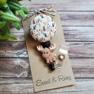 Pu&ograve; includere: Un portabadge con un bottone circolare in tessuto con motivo ad alberi, un ciondolo in silicone a forma di alce marrone e un cinturino in plastica trasparente con bottone a pressione. Fissato su un cartoncino marrone con scritta Scout & Remy.