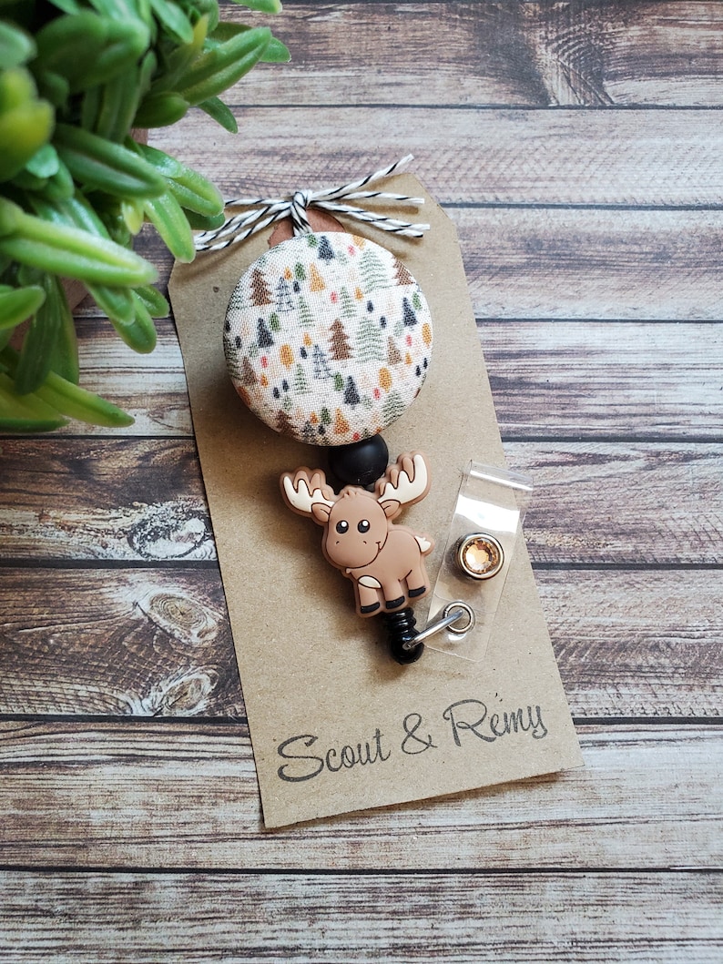 Pu&ograve; includere: Un portabadge con un bottone circolare in tessuto con motivo ad alberi, un ciondolo in silicone a forma di alce marrone e un cinturino in plastica trasparente con bottone a pressione. Fissato su un cartoncino marrone con scritta Scout & Remy.