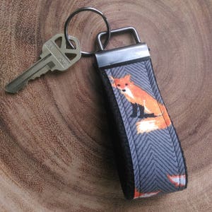 May include: A gray and orange fox keychain with a black metal key ring and a silver key. The keychain is on a wooden surface. "SCOUT AND REMY" is printed on the surface.