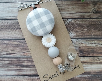 Può includere: Un portabadge con un bottone a quadretti grigio e bianco, una margherita, perline grigie e di legno e il testo "Scout & Remy" su un'etichetta marrone. L'avvolgitore è attaccato a una clip di plastica trasparente.