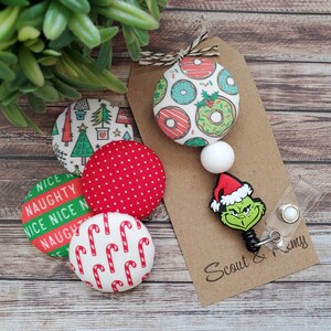 May include: A set of five Christmas-themed fabric buttons with a Grinch-themed retractable badge holder. The buttons feature red and white candy canes, red polka dots, green and white Christmas trees, and green and white doughnuts. The badge holder has a white bead, a green Grinch face, and a red Santa hat. The text on the badge holder reads "Scout & Remy".