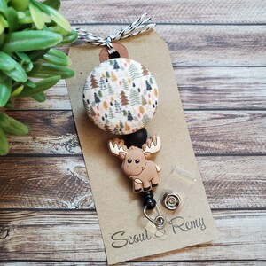Pu&ograve; includere: Un portabadge con un bottone circolare in tessuto con motivo ad alberi di bosco multicolore, un ciondolo in silicone marrone a forma di alce e una perlina nera. L'articolo &egrave; fissato su un cartoncino marrone con il testo Scout & Remy.