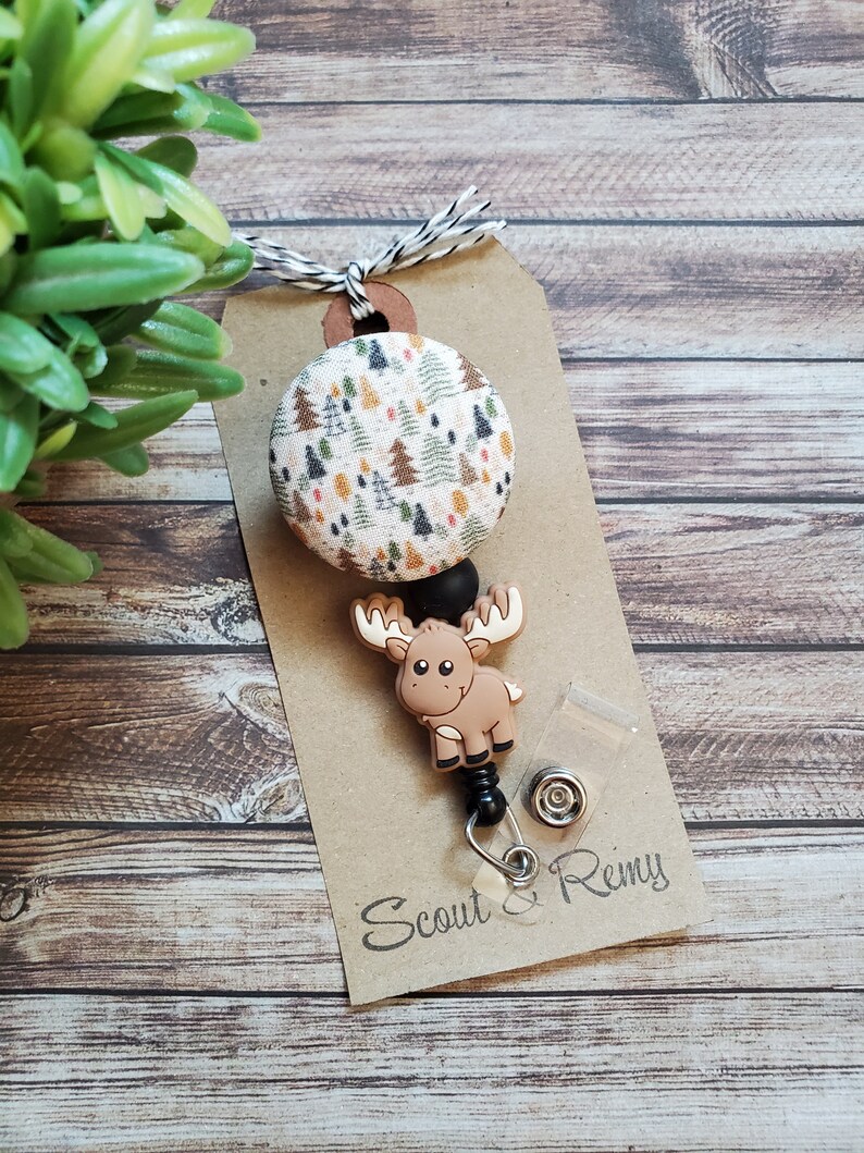 Pu&ograve; includere: Un portabadge con un bottone circolare in tessuto con motivo ad alberi di bosco multicolore, un ciondolo in silicone marrone a forma di alce e una perlina nera. L'articolo &egrave; fissato su un cartoncino marrone con il testo Scout & Remy.