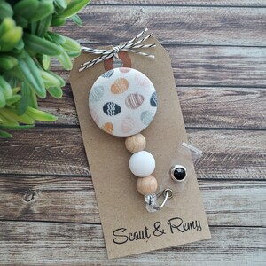 May include: A white and brown beaded mask chain with a black button and a round fabric charm with a colorful egg pattern. The charm is attached to a brown card with the text "Scout & Remy".