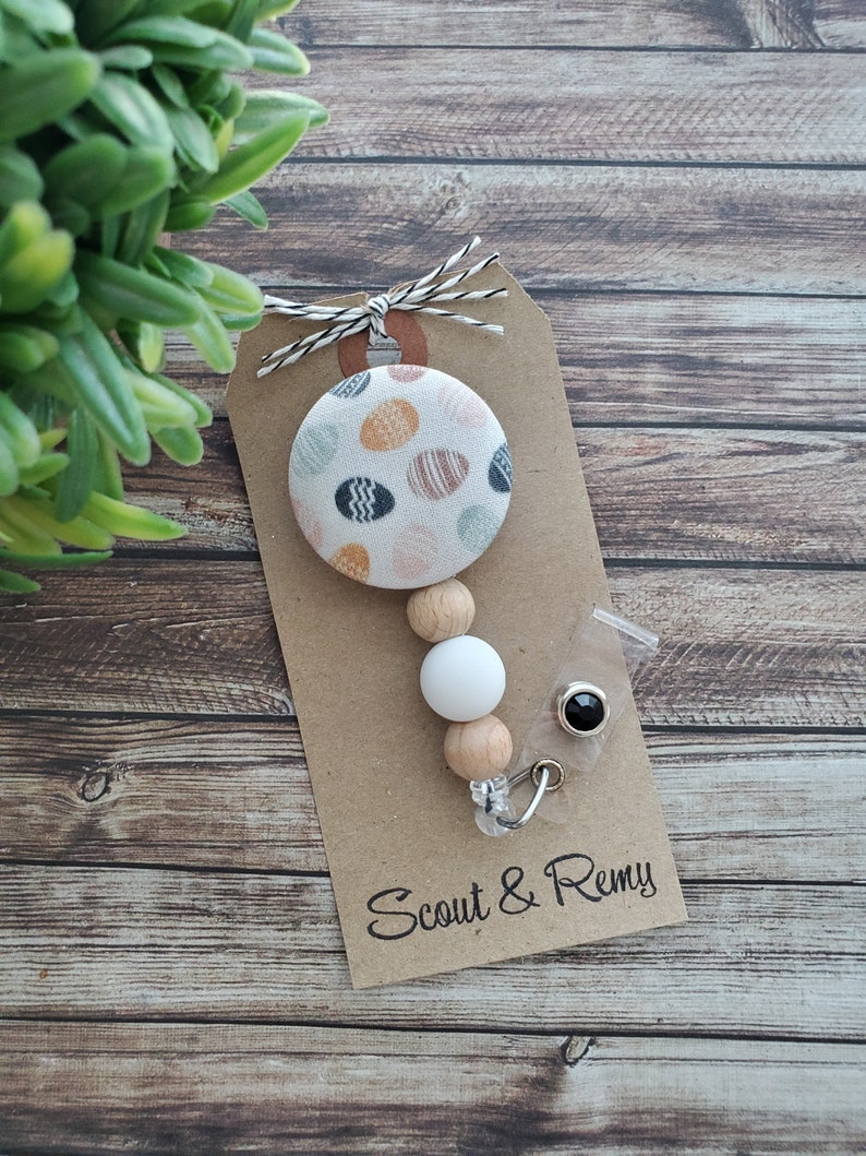 May include: A white and brown beaded mask chain with a black button and a round fabric charm with a colorful egg pattern. The charm is attached to a brown card with the text "Scout & Remy".