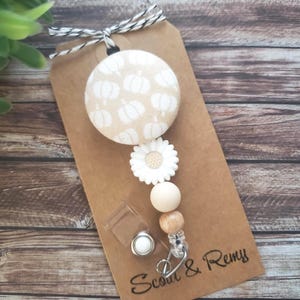 May include: A beige and white pumpkin-themed badge reel with a white daisy accent and wooden beads. The reel is attached to a brown tag with the text "Scout & Remy" and a black and white twine bow.