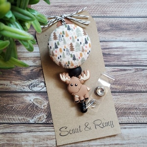 Pu&ograve; includere: Un portabadge con un bottone circolare in tessuto con motivo ad alberi di bosco multicolore, un ciondolo in silicone marrone a forma di alce e una clip in plastica trasparente. L'articolo &egrave; fissato su un cartoncino marrone con la scritta Scout & Remy.