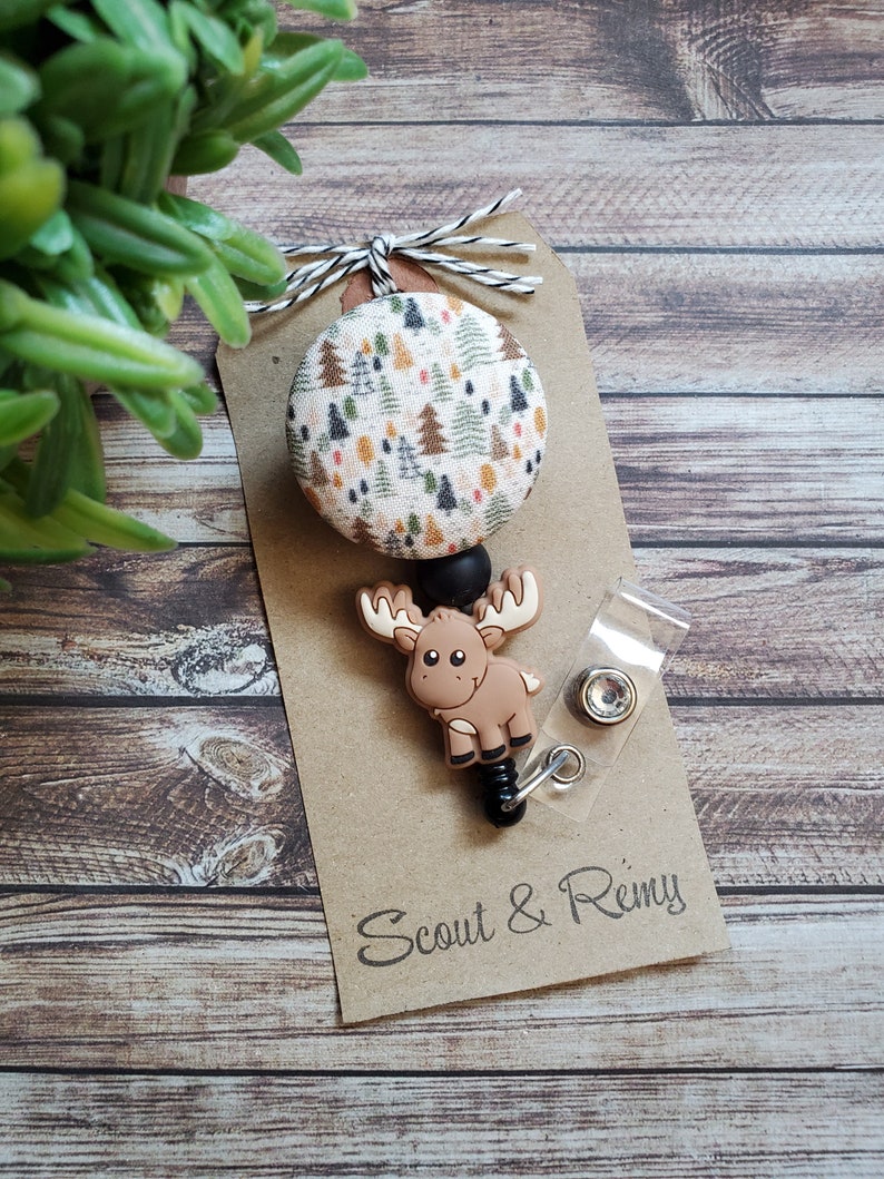 Pu&ograve; includere: Un portabadge con un bottone circolare in tessuto con motivo ad alberi di bosco multicolore, un ciondolo in silicone marrone a forma di alce e una clip in plastica trasparente. L'articolo &egrave; fissato su un cartoncino marrone con la scritta Scout & Remy.
