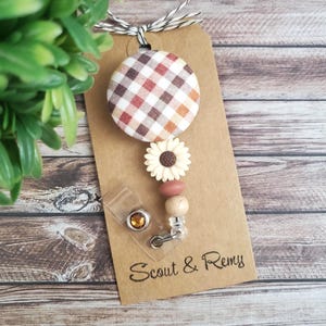 May include: A badge reel with a plaid fabric button, a daisy accent, and wooden beads. The reel is attached to a brown tag with the text "Scout & Remy". The reel is attached to a brown tag with the text "Scout & Remy".