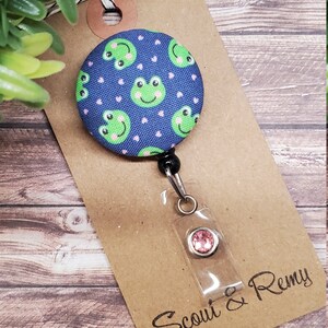 Puede incluir: Un carrete de insignia de tela azul y rosa con un patr&oacute;n de rana verde y un strass rosa. El carrete est&aacute; unido a una tarjeta marr&oacute;n con el texto "Scout & Remy".