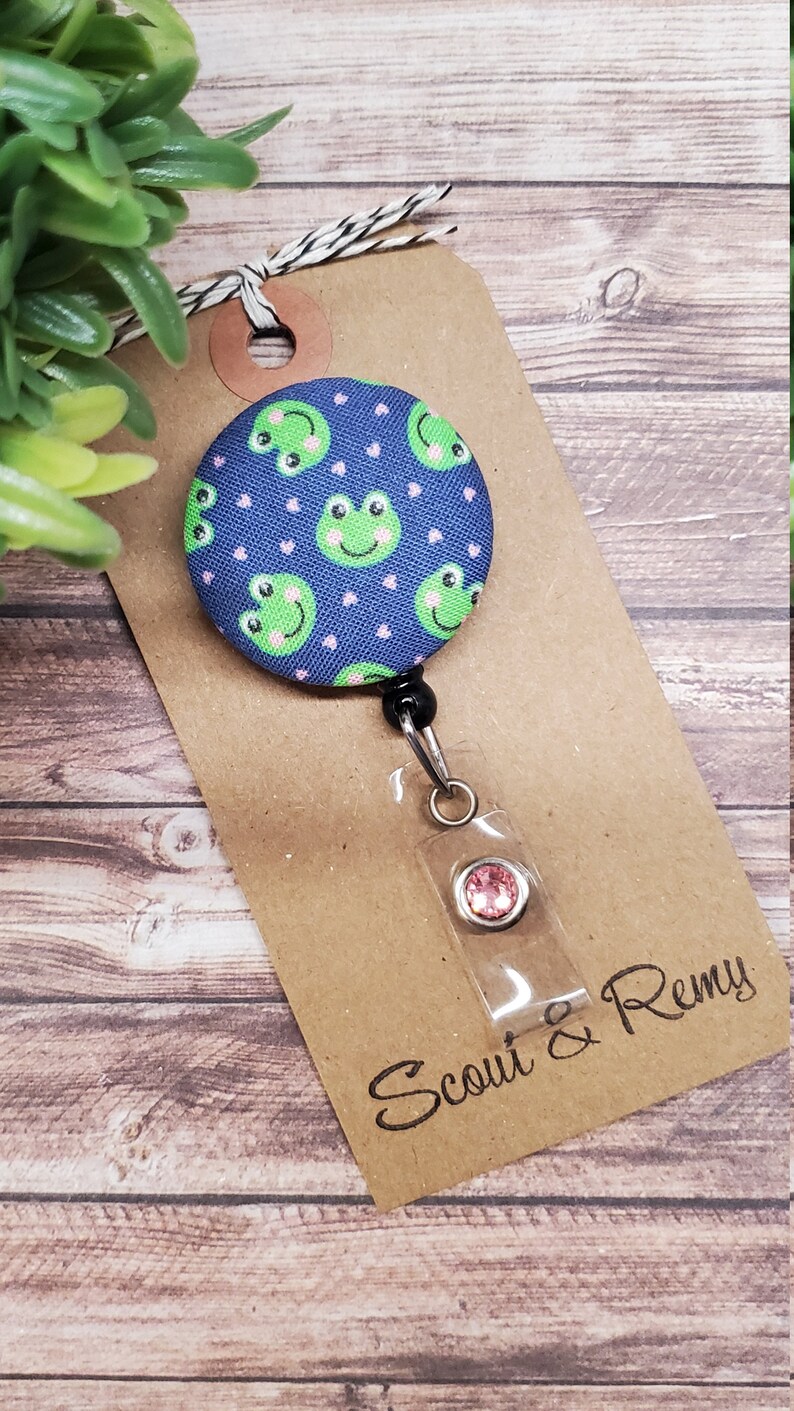 Puede incluir: Un carrete de insignia de tela azul y rosa con un patr&oacute;n de rana verde y un strass rosa. El carrete est&aacute; unido a una tarjeta marr&oacute;n con el texto "Scout & Remy".