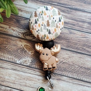 Pu&ograve; includere: Un portabadge retrattile con un bottone circolare in tessuto caratterizzato da una stampa boschiva con piccoli alberi verdi, marroni e arancioni. Sotto il bottone si trova un ciondolo a forma di alce in silicone marrone, un connettore nero e una cinghia.