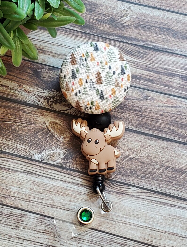 Pu&ograve; includere: Un portabadge retrattile con un bottone circolare in tessuto caratterizzato da una stampa boschiva con piccoli alberi verdi, marroni e arancioni. Sotto il bottone si trova un ciondolo a forma di alce in silicone marrone, un connettore nero e una cinghia.