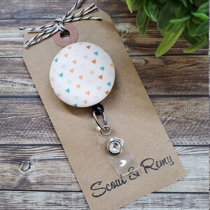 May include: A white fabric badge reel with a colorful heart pattern and a clear plastic badge holder. The reel is attached to a brown card with the text "Scout & Remy".