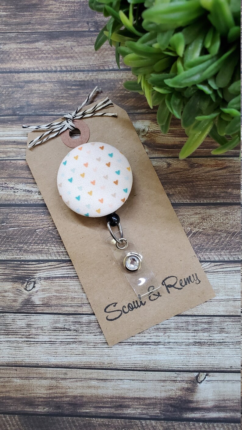 May include: A white fabric badge reel with a colorful heart pattern and a clear plastic badge holder. The reel is attached to a brown card with the text "Scout & Remy".