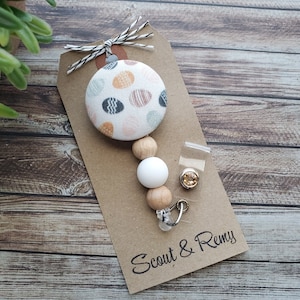 May include: A brown card with the text "Scout & Remy" printed on it. A white and brown beaded retractable badge holder with a fabric button featuring a white background with colorful polka dots.