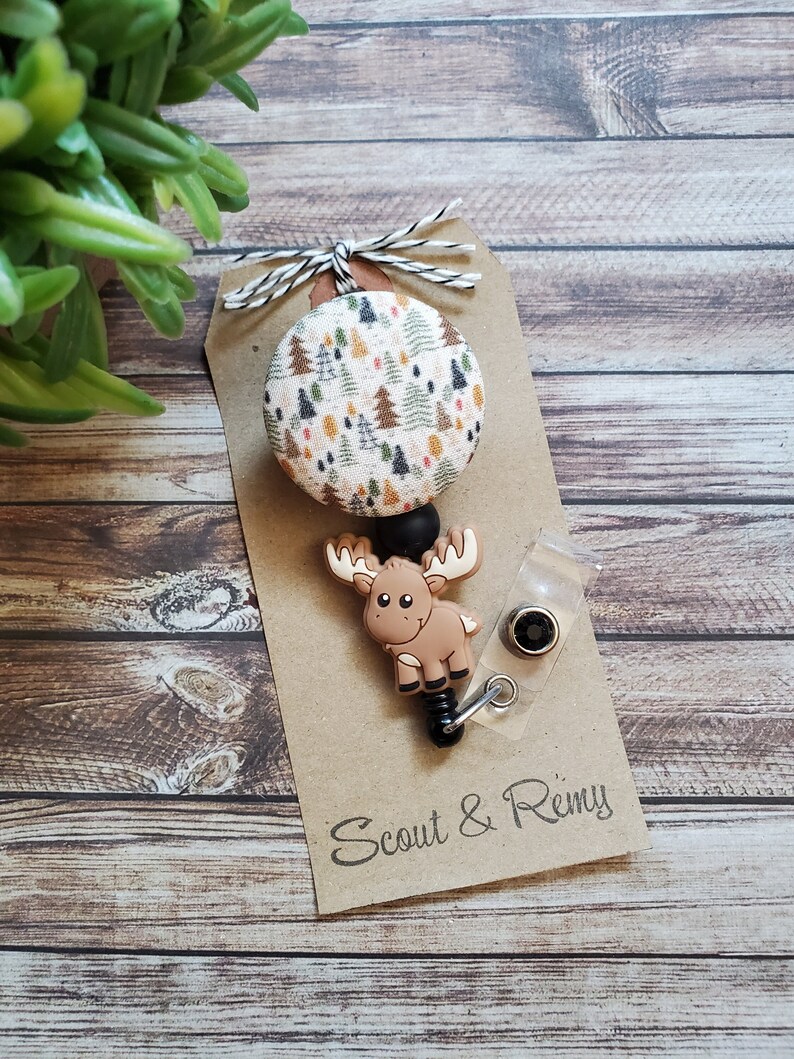 Pu&ograve; includere: Un portabadge retrattile con un bottone circolare in tessuto con motivo di alberi di bosco multicolore e un ciondolo in silicone a forma di alce marrone. Il portabadge &egrave; fissato su un cartoncino marrone con il testo Scout & Remy e un fiocco.