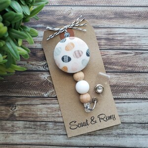 May include: A white and brown beaded phone charm with a colorful patterned button and a clear crystal charm. The charm is attached to a brown card with the text "Scout & Remy".