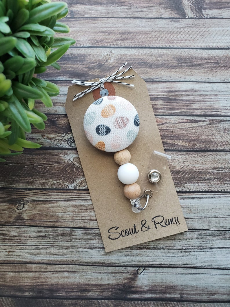 May include: A white and brown beaded phone charm with a colorful patterned button and a clear crystal charm. The charm is attached to a brown card with the text "Scout & Remy".